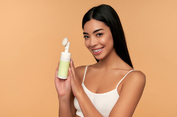 Young asian woman with radiant skin cleans her face with a brush and foam.isolated on a beige background. Facial skin care concept. Beauty.