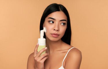 Obraz na płótnie Canvas Young asian woman with radiant skin cleans her face with a brush and foam.isolated on a beige background. Facial skin care concept. Beauty.