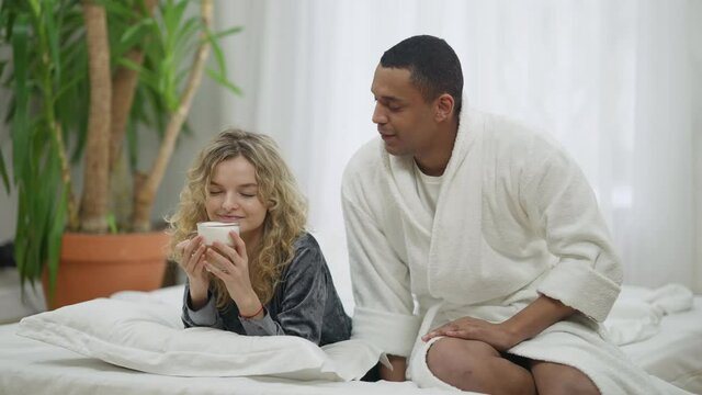 African American loving man bringing morning coffee for Caucasian woman in bed. Wide shot portrait of happy smiling boyfriend taking care of girlfriend in bedroom at home. Love and romance