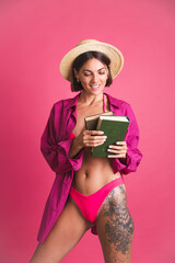 Beautiful fit tanned sporty woman in bikini and shirt holding book go to beach to read on pink background