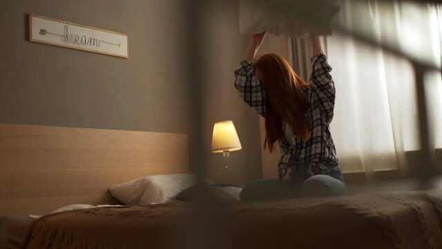 Angry disgruntled young woman screaming into pillow and hits bed with pillow in bedroom on background of window. Mad crazy redhead female vented anger in privacy of home. Shooting in slow motion.