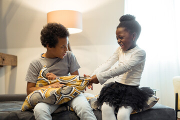 Two African American ethnic kids are playing on the sofa and beating each other with pillows.