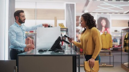 Clothing Store: Young Woman at Counter Buys Clothes from Friendly Retail Sales Assistant, Paying with Contactless NFC Smartphone Touching Terminal. Trendy Fashion Shop with Designer Brands. - Powered by Adobe