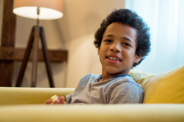 Fototapeta premium A cute little boy with an afro hairstyle sits comfortably on the sofa and looks at the camera.