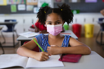 Portrait of african american girl wearing face mask sitting on her desk in the class at school