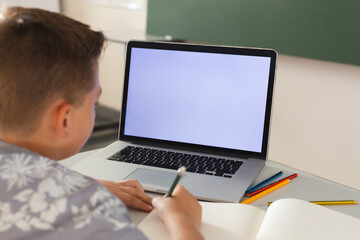 Fototapeta na wymiar Caucasian boy sitting at a desk in classroom writing and using laptop, with copy space on screen