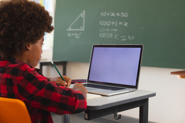 African american schoolboy sitting at desk in classroom using laptop, with copy space on screen