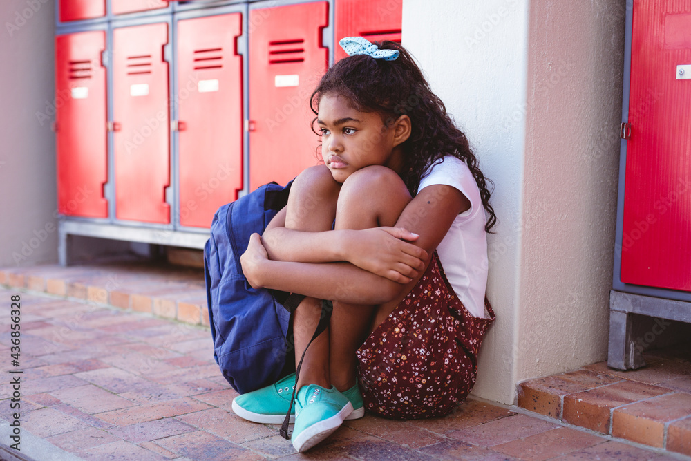 Wall mural Unhappy african american schoolgirl sitting by lockers in school corridor with schoolbag