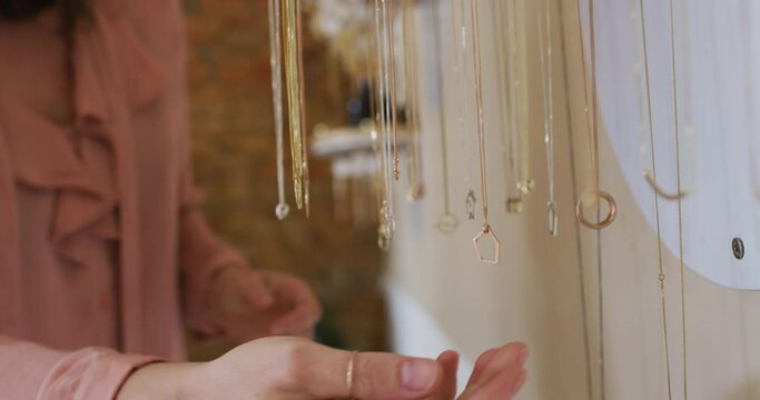Close up of hand of caucasian woman touching golden necklaces hanging on rack, shopping for jewelry