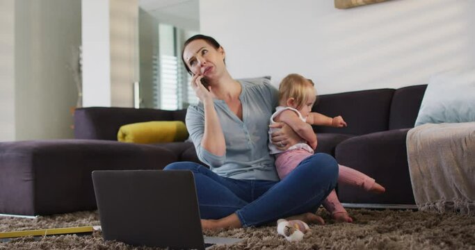 Caucasian mother holding her baby talking on smartphone while working from home