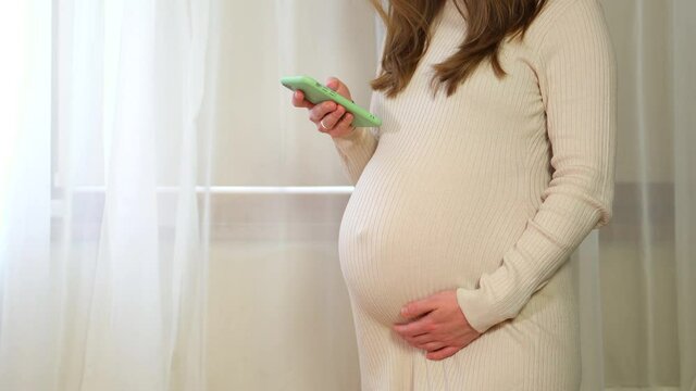 pregnant woman holds phone and calls doctor, watches news, scrolls messages, plays online. Pregnancy and motherhood concept. Close-up of pregnant woman belly with unborn baby and smartphone in hands.