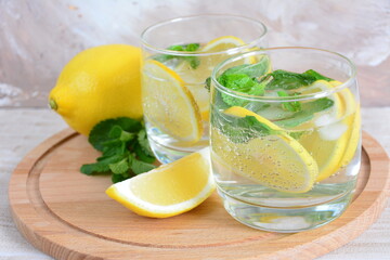 glasses with mineral water, lemon slices, mint leaves and ice cubes on the cutting board on the beige background