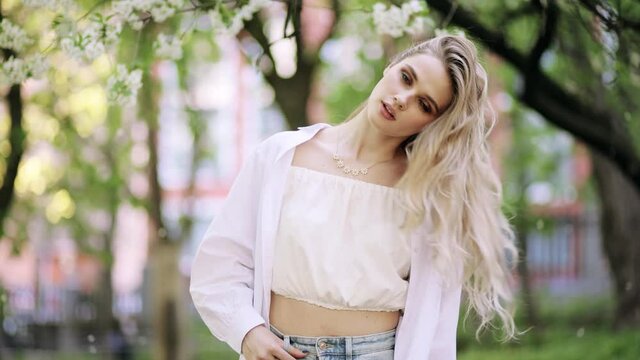 Attractive fashion model with flying hair in the flowered garden in the spring time. Young beautiful blonde woman shakes hair. Blossoms fall from trees. Slow motion