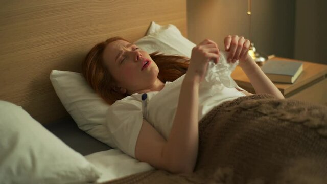 Close-up of sick young woman sneezing, blowing and wiping nose with napkin lying in bed. Closeup of Ill female checking high temperature with thermometer, suffering from flu or coronavirus.