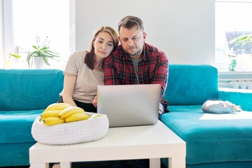 Serious middle-aged couple husband and wife with laptop sitting at home on couch