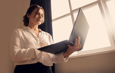 Middle-aged business woman stands in the office near the window, holding open laptop