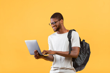 Smart african american guy with backpack using laptop for online distant work or studies over yellow background