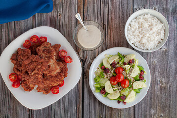 a plate with breaded chicken with cherry tomatoes, salad and a small glass bowl of dressing or gravy ,with a blue napkin on th side, over a rustic table