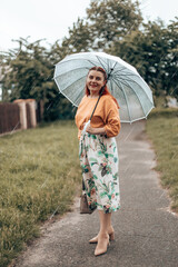 Happy young woman in bright clothes under a transparent umbrella in bad weather in the city park