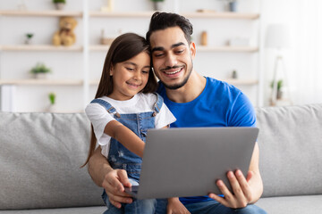 Smiling dad and daughter sitting on couch, using pc