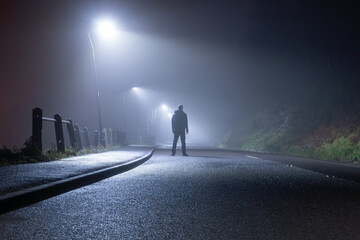 A mysterious man, alone, standing in the middle of a country road. Under street lights. On a foggy,...