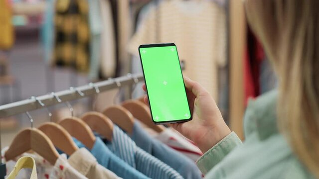 Clothing Store: Female Using Smartphone with Chroma Key Green Screen Display. Clothes Hanger with Stylish Branded Items for Retail Sale In the Background. Close Up Footage of Mobile Device.
