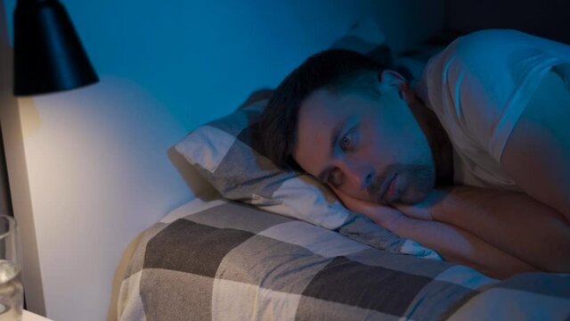 Stressed man trying to sleep in bed at night. Young male with open eyes suffers from insomnia and sleep disorders. Things dont feel same anymore. Blue tones. Sleepless man. Guy dont sleep at night