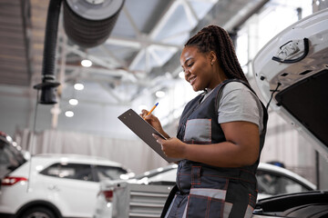 Afro american Woman Mechanic writing on clipboard at repair garage, wearing uniform overalls. Young...
