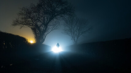 A scary, mysterious hooded figure, standing in front of a bright light on a country lane, on a...