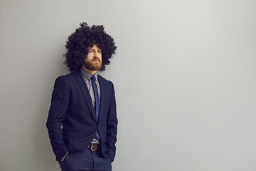 Young businessman with curly hair looking away and thinking standing hands in pockets on grey copy space background. Serious pensive man with funny hairstyle leaning on copyspace studio wall