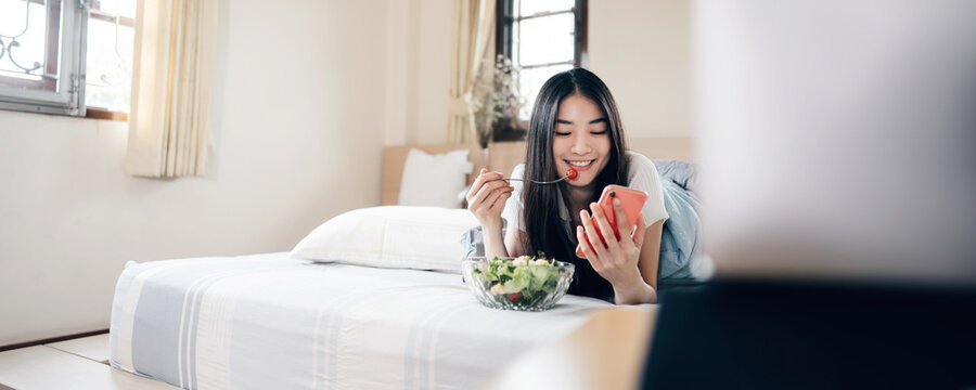 Young adult asian woman eating salad and using mobile phone and laptop computer for watching online movie on internet in bedroom.