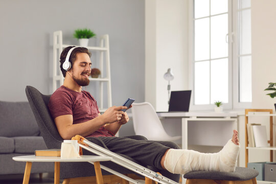 Positive man enjoying sick leave despite physical injury. Happy smiling young guy with broken leg in plaster cast sitting in chair at home, using mobile phone and listening to music through headphones