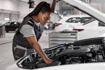 Afro Car mechanic woman is examining under hood of car at repair garage, wearing overalls, looking...