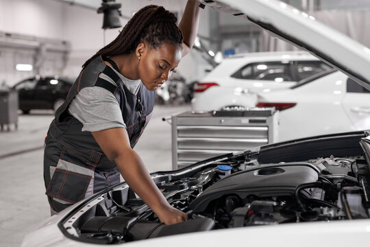 Afro Car Mechanic Woman Is Examining Under Hood Of Car At Repair Garage, Wearing Overalls, Looking Confident And Concentrated. Side View On Female Trying To Solve The Problem Of Inoperative Engine