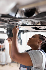 Black Female Auto Mechanic Working Underneath Car In Garage. Portrait of hardworking young female mechanic in overalls, inspecting lifted car. Mechanic woman working in car repair shop. Side View