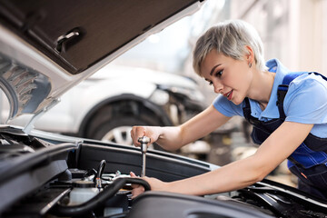 Fototapeta na wymiar Focused Female Professional Mechanic Working on Vehicle in Car Service. Short Haired Female Engine Specialist Fixing Motor, Wearing Overalls and Using a Ratchet. Modern Clean Workshop