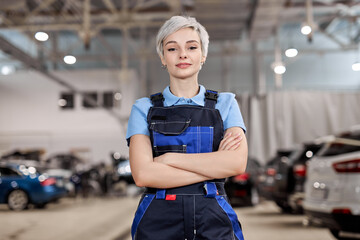Caucasian professional female mechanic posing at camera standing in auto repair shop. Short haired woman in blue uniform. Car service, repair, maintenance and people concept. Front view. Copy space - obrazy, fototapety, plakaty