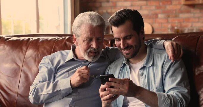 Millennial man sit on sofa hold smartphone showing to older dad new mobile application, explain usage of most useful medical apps for seniors, discussing media news. Modern tech and elderlies concept