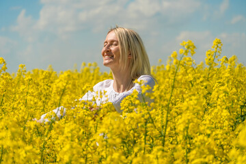 Naklejka na ściany i meble Portrait of a beautiful happy young blonde woman in a white shirt in a rapeseed field on a sunny day