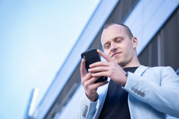 Bottom view perspective of young short-haired businessman in a blue suit standing in front of a corporate building and writes a text message on mobile.