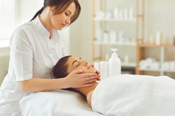 Obraz na płótnie Canvas Smiling doctor dermatologist making procedure of relaxing facial massage for young woman patient in beauty spa salon. Rejuvenating and lifting massage in cosmetology concept