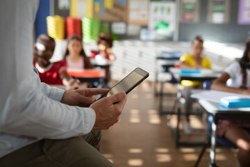 Mid section of male teacher using digital tablet while sitting in the class at elementary school