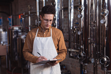 Caucasian male worker wearing apron writing on clipboard at gin distillery
