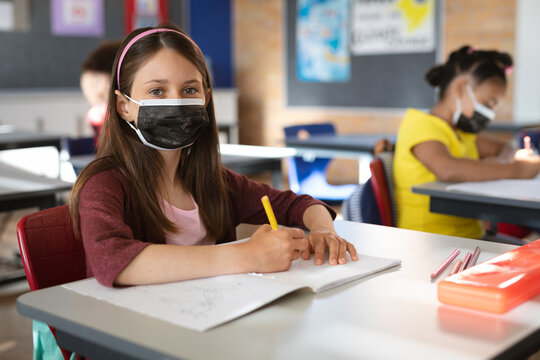 Portrait Of Caucasian Girl Wearing Face Mask Sitting On Her Desk In The Class At Elementary School