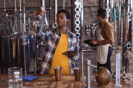 African american female worker holding a flask and digital tablet at gin distillery