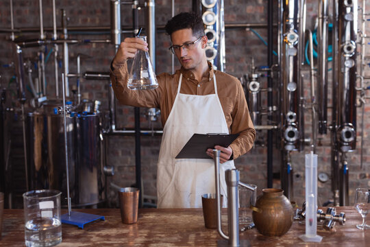 Caucasian male worker wearing apron holding a flask and clipboard at gin distillery - Powered by Adobe