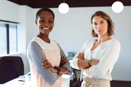 Two diverse smiling businesswomen standing with arms crossed at work