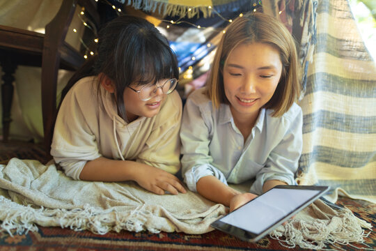 Smiling asian woman with her daughter using tablet lying under tent in living room
