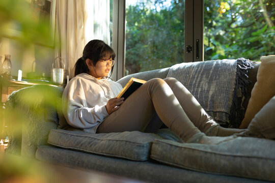 Smiling asian girl in glasses reading a book and sitting on sofa