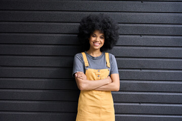 Young beautiful black hipster girl of 20s standing with crossed arms on black wall background....